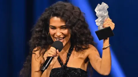 Olivia Dean wears a black sparkly strappy dress. She is dark brown hair and wears hoop earings. She is standing on stage accepting a silver trophy.