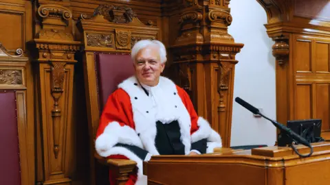 Bailiff of Jersey Robert MacRae in a chamber. He is sitting on a wooden seat which is lined with leather and modeled a little like a throne. He is smiling at the camera and is wearing a red robe with white ermine trim over a black suit. 