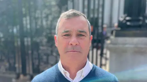 Alan Brecknell has short greying hair and is wearing a blue v-neck jumper with an open necked white shirt