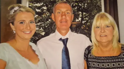 Family handout Three people stand smiling for the camera in front of a window, which shows large trees outside. To the left is a young woman in a green dress. Her blonde hair is tied up in a bun with a floral display. She has white earrings dangling. A man in a white shirt and blue tie is in the centre with an arm around the two women. He has dark hair, which is turning grey. To the right is an older woman with blonde hair which hangs by her shoulders. She is wearing a black pattered top, which appears to have flowers embroidered into the pattern.
