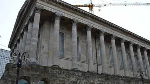 Birmingham town hall, a brick building with huge stone pillars around it. There is a brick wall with archways beneath it. A yellow crane is behind it, looming above
