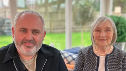 Justin, a middle aged man with a beard and black zip up top, sitting next to Mary, an older woman with grey hair and a patterned top. Behind them are large windows looking out to a garden with a grass lawn.