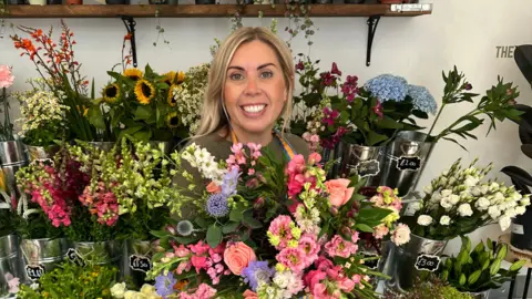 Charlotte Phipps A blonde haired woman smiling while surrounded by pink, purple and yellow flowers in a florist shop.