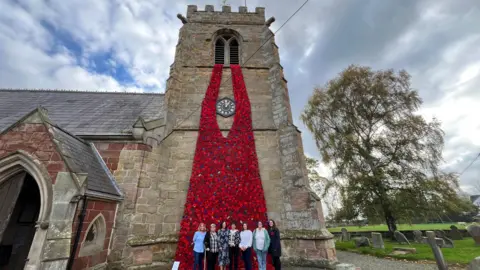 ELLEN KNIGHT/BBC A stone church, with a huge red cascade of knitted poppies 'waterfalling' out of the windows in the belfry all the way down to the ground. In front of the cascade are stood seven women side by side. The sky is blue and cloudy with a bit of sunshine. 