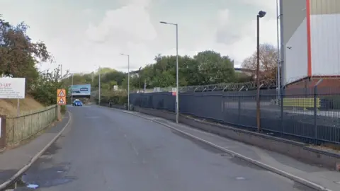 A car is driving along a road lined with trees and an industrial estate to the right.