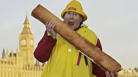 The late Grimsby MP Austin Mitchell pictured with a giant fish finger, superimposed in front of the Houses of Parliament. Mr Mitchell is wearing a red and yellow fisherman's outfit, including a yellow hat with "Grimsby" emblazened on the front. Big Ben can be seen in the background. 