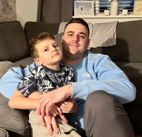 Danny Buckley and his son Arlo sit cuddled together on a grey sofa, looking at the camera.