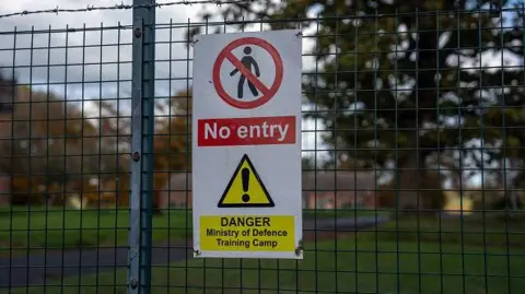 Getty Images A warning sign reading "No entry" and "Danger Ministry of Defence Training Camp" is attached to a wire fence topped with barbed wire. Open grassland can be seen beyond the fence.