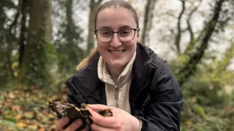 BBC Niamh Carmichael, a woman with fair hair, glasses, dark jacket and brown fleece, holding leaves in her hands.