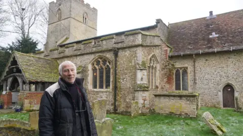 Diocese of St Edmundsbury and Ipswich Churchwarden Peter Thompson, wearing a dark jumper and jacket, stands in the grounds of St Margaret's Church in Stradishall, with the entrance, side of the church and tower visible behind him.