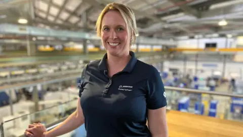BBC/Laura Jones A woman with short blond hair is smiling and wearing a black polo shirt with the word Boeing in white letters. She is standing on a mezzanine floor overlooking a factory