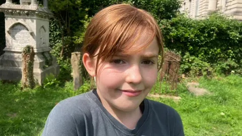 A 10-year old girl is standing in an old graveyard. She is ginger hair with a fringe, and is wearing a grey- shirt. 
