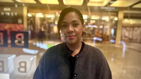 A woman in a dark grey cardigan looking into the camera with a small smile and has her hair up. Part of her hair is hanging over her face and she has red lip gloss on. 