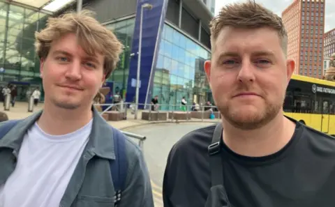 Michelle Adamson/BBC Ben Esson (left) has short wavy brown hair and a goatee and wears a grey jacket and white T-shirt. Callum KIrk has dark brown hair and a beard and wears a black T-shirt. They stand outside Manchester Piccadilly Station.