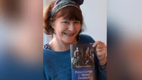 Rosalind Freeborn is wearing a blue jumper and is smiling at the camera. She is holding a copy of her book, Prince George and Master Frederick.