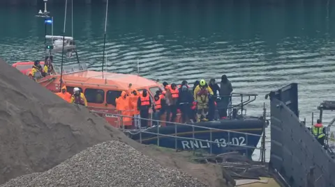 A group of people thought to be migrants are brought in to Dover, Kent, by the RNLI Dungeness Lifeboat following a small boat incident in the Channel.  They are wearing red life jackets and standing on the bow of the lifeboat.