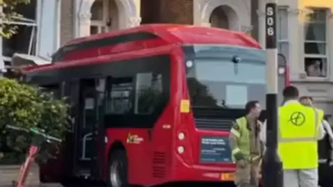 A red bus is crashed into the front of a building. People stand behind it.