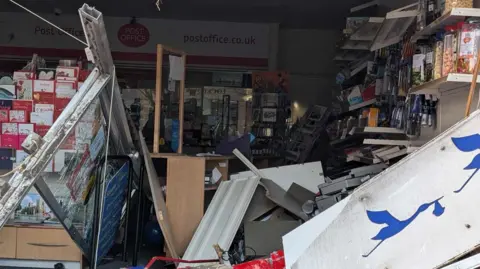 Sarah Barnett Close up image showing the damage to the post office in Winterbourne. A stand of greeting cards can be seen to the left of the frame and there is a lot of damaged stock and fixtures from the inside of the post office on the floor, blocking the aisle.