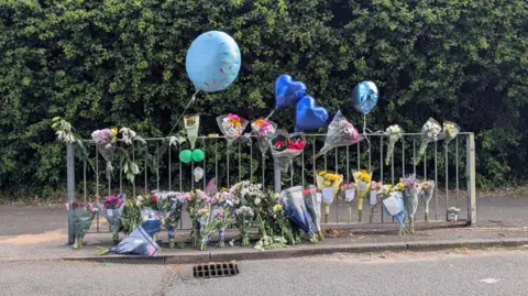 Flowers and balloons next to a metal barrier on the side of the road.