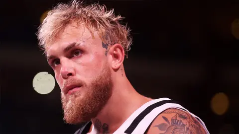 Jake Paul looking to the left, wearing a white and black vest. A tattoo on the side of his head is visible, and the background is black.