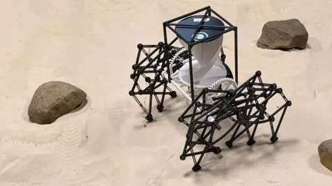 A robotic creation comprising a black plastic cuboid frame, raised off the ground by two black plastic geometric shapes attached by white plastic cogs. The structure is pictured on sand, beside three small brown rocks 