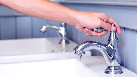 Getty Images A man holding the handle of a chrome tap attached to a sink. Another chrome tap and sink are in the background. Blue tiling surrounds the taps and sinks.