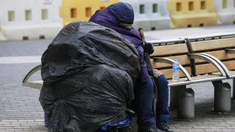 PA Media File image of a homeless person resting on a bench next to a pile of belongings covered by black sacks