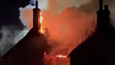 Orange flames are burning intensely in the roofs and chimney of a cottage on the left of the shot and appears to have spread to the next door home.