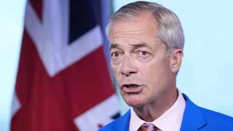 Nigel Farage in a blue suit and pink shirt and tie, standing in front of a blue background with a union flag in the background