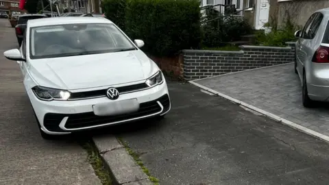 A white Volkswagen car parked on a pavement, slightly blocking a neighbours' grey brick driveway. A silver Volkswagen is parked on the driveway in front of a row of terraced houses. 