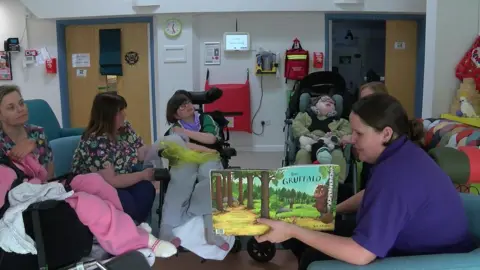 A woman in a purple t-shirt working at the hospice reading The Gruffalo to a crowd of children who are sat around in specialist chairs listening. They are each being supported by adults.