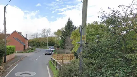 A residential street with a wooden pole on the right and bushes and cars on the right of the road and a red brick house on the left.