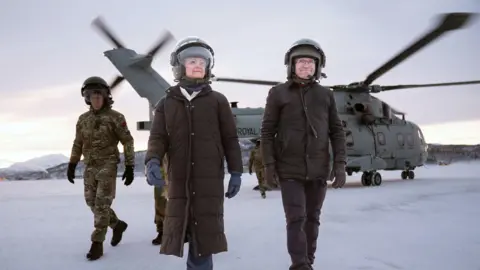 PA Media A woman dressed in a dark brown coat, navy blue gloves, blue trousers, brown boots and a helmet walks alongside a man in dark brown trousers, a dark brown jacket, boots and a helmet. Behind them, soldiers wearing camo unfiform disembark from a helicopter. The tarmac is snowy and the sky is grey.