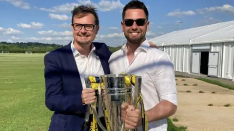 Oxford United chief exec Tim Williams, a white middle aged man with brown hair wearing a blue blazer, white shirt and black framed glasses, stands with head coach Des Buckingham, a white middle aged man with dark short spiked hair in a white shirt and sunglasses holding the trophy