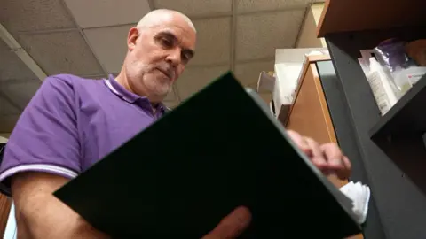 A middle-aged man with shaved white hair and beard, wearing a purple polo shirt, reads from a black ring-binder.