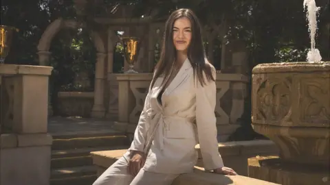 Vanesa, with long brown hair and wearing a cream suit