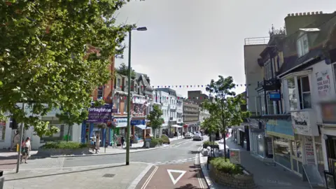 A road junction with a number of shops either side of a road with trees either side of the road.