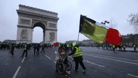 AFP A demonstrator in a wheelchair is pushed by another, holding a French national flag with a yellow one attached to it, with the Arc de triomphe in the background during a Yellow Vests "Gilets Jaunes" anti-government demonstration in Paris on 12 January 2019