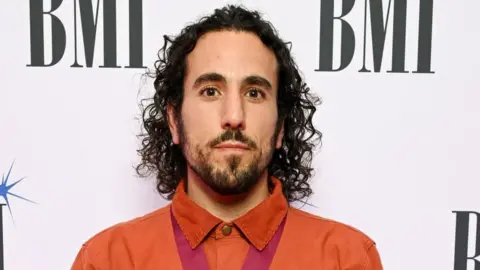 Getty Images Aaron Horn at the BMI Awards. Aaron has curly dark hair cut just above his shoulders and a short dark beard. He has brown eyes and looks at the camera. He is wearing a burnt orange shirt buttoned to the top with a red silk around his neck which has a medal on it. He is pictured in front of a white back drop with the BMI logo printed on it.