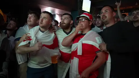 Five England football fans gathered in The Hyde Dendy pub in Paignton to watch the game against Mexico. They are all staring intently at the TV, all dressed in England red and white colours. 