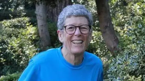 Submitted A woman with tightly curled short, grey hair smiles into the camera. She wears a blue t shirt and black, round glasses.