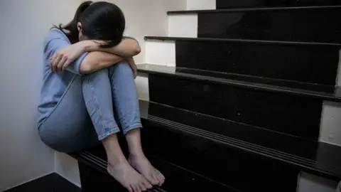 Getty A barefoot woman wearing light blue jeans and a light blue top is sitting down on some stairs with her arms around her knees and her face in her lap. she is pale with dark brown hair in a ponytail