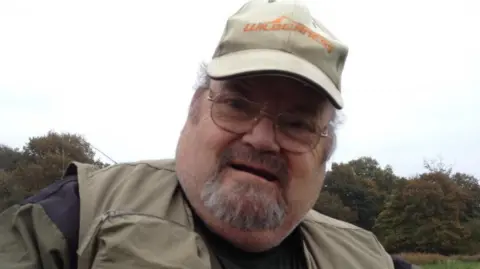 Alan Graham pictured holding a fish. He is wearing a khaki hat and fisherman's jacket. He has a grey beard and wears circular silver framed glasses