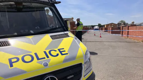 The front of a police van which is parked nearby a police cordon with a police officer. A white forensic tent can be seen in the background.