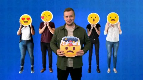 A man stands in the middle of a group of four people holding an emoji in his hand. The rest of the group have emojis for faces