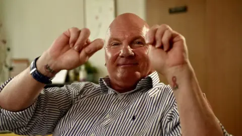 Ed Hanson/BBC Nigel Bullock has a bald head and wears a black and white striped button-up shirt. He is sitting down smiling at the camera with his arms raised in front of him. He has a small tattoo on the pulse point of each wrist - one showing the HIV awareness symbols and the other with the symbol U=U. He is wearing a black leather-strapped watch. He is in an office.