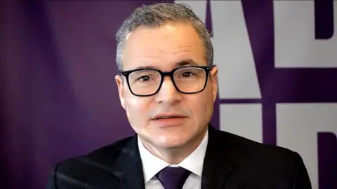 ADHD UK Henry Shelford has short grey hair and black glasses. He is wearing a black suit and a purple tie.