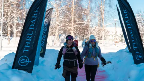 Beyond the Ultimate A woman in black arctic gear with black hiking poles is smiling. The hood is up and she has a pink neck scarf around her chin. There is a pink rucksack on her back with water bottles on the straps. She is smiling at a woman next to her who is wearing a grey ski jacket, black trousers and a large rucksack, with a grey beanie and blue sunglasses. They are sunning down a pink carpet which is surrounded by snow, with snowy trees in the background. the sun is shining