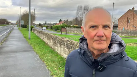 Fairburn parish councillor John Kos is looking straight at the camera. He is wearing a blue puffa-style jacket and has thinning, grey hair.