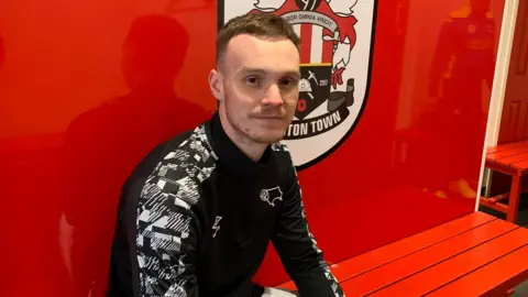 Harry Cook, he is sat on a red bench, wearing a black tracksuit top